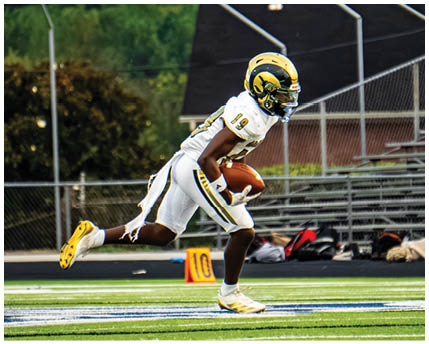 A person wearing a green and white football uniform with a gold helmet is bent forward on a field, preparing for a play. Description generated by AI