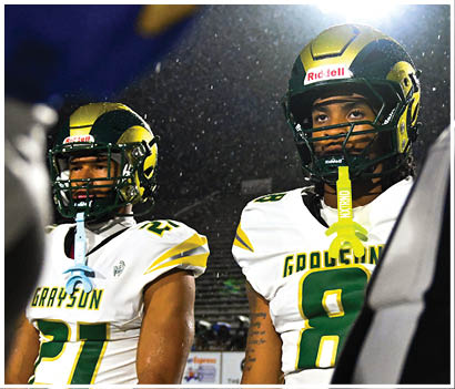 Two people in white and green football uniforms stand on a field with one clenching a fist and a referee and opposing players in red uniforms visible in the background. Description generated by AI