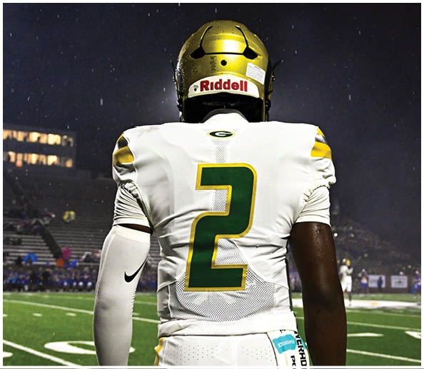 A person in a white and gold football uniform is crouched in an athletic stance on a green football field with other players and a coach in the background. Description generated by AI