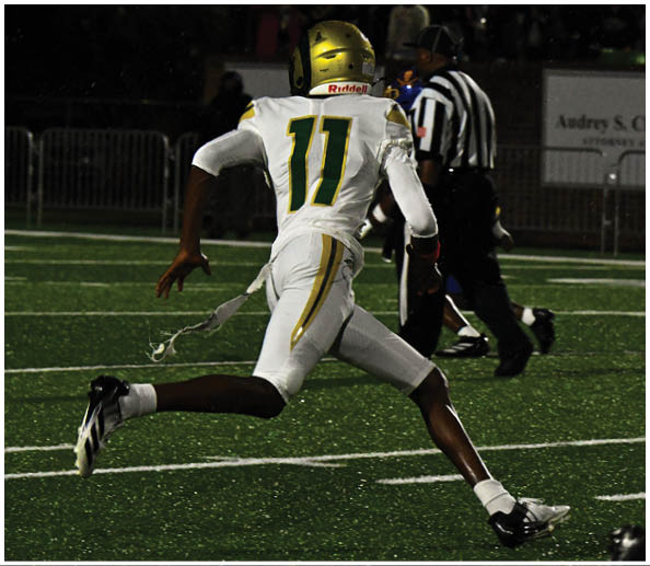 A person in a white and green football uniform with a gold helmet is on a field facing away from the camera while another person in a maroon and gold uniform stands in the background near empty bleachers. Description generated by AI