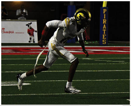 A person wearing a white and gold football uniform with the number 20 stands on a field while two other people in red and gold football uniforms are visible in the background near empty bleachers. Description generated by AI