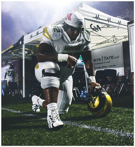 Two people in football uniforms celebrating on a field with a chain-link fence and a blue Academy Sports + Outdoors banner in the background. Description generated by AI