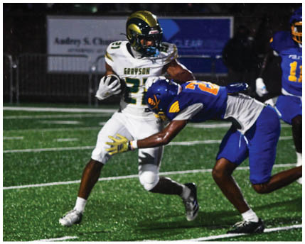 Three people wearing white and gold football uniforms and helmets are standing and interacting near a chain-link fence with trees and bleachers in the background. Description generated by AI