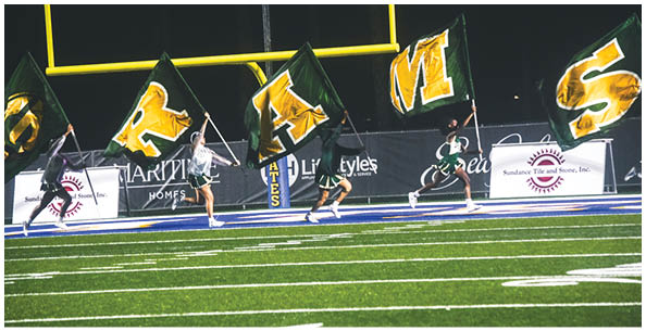 Four people wearing white football uniforms with gold helmets and large numbers on their backs are standing on a field. Description generated by AI