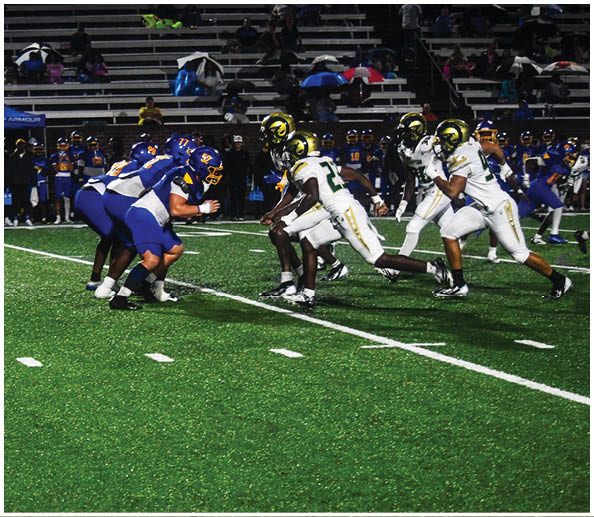 A person in a white football uniform with the number 12 and a gold helmet stands on a football field with blurred spectators and cheerleaders in the background. Description generated by AI