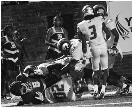 A person in a white and yellow football uniform is crouched on a green field with a referee and other players in the background. Description generated by AI