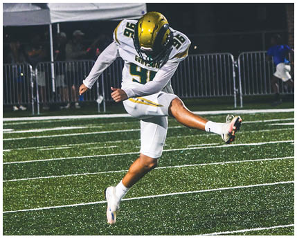 A person wearing a white football uniform with a gold and black helmet is kneeling on a green football field with arms extended forward. Description generated by AI