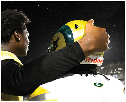 Two people wearing football helmets and white jerseys with gold and green accents are facing each other in a stadium with empty bleachers in the background. Description generated by AI