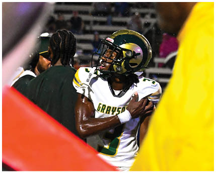 A person wearing sunglasses and a headset is speaking intensely while another person in a football helmet looks on in the background. Description generated by AI