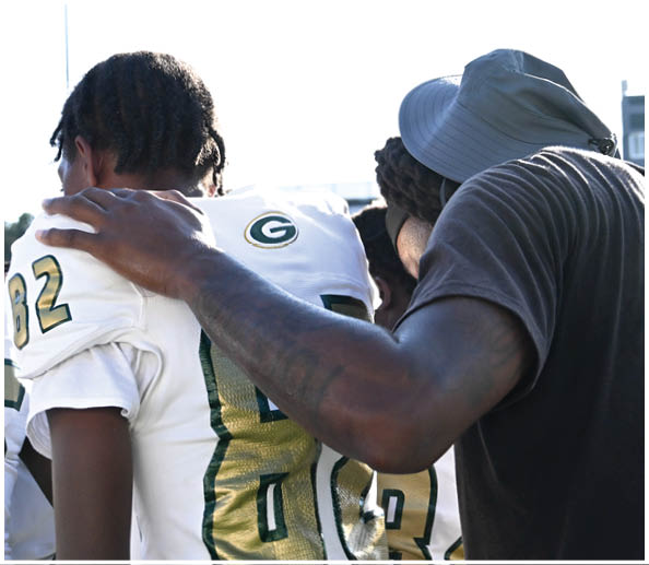 A person in a white and green football uniform with a gold helmet is on a field facing away from the camera while another person in a maroon and gold uniform stands in the background near empty bleachers. Description generated by AI
