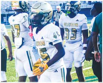 A person wearing a white and gold football uniform with the number 20 stands on a field while two other people in red and gold football uniforms are visible in the background near empty bleachers. Description generated by AI