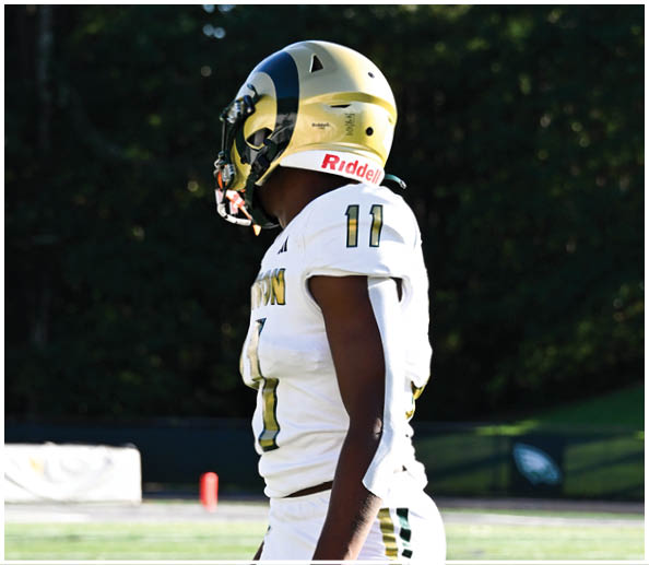 A person in a white and gold football uniform is sitting on the field holding a football while other players stand in the background. Description generated by AI