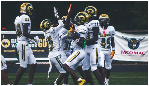 A person wearing a white football uniform and helmet is running with a football on a field while people in green cheerleading outfits stand in the background. Description generated by AI