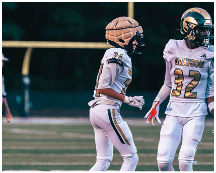 A person wearing a white football uniform with a gold and black helmet is kneeling on a green football field with arms extended forward. Description generated by AI