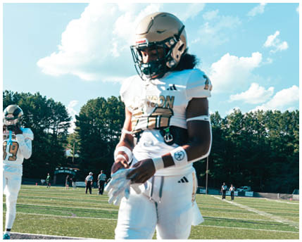 A person in a white and gold football uniform is crouched on a field with other players in similar uniforms standing in the background. Description generated by AI