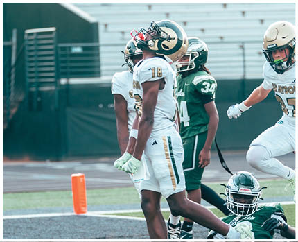 Two people wearing football helmets and white jerseys with gold and green accents are facing each other in a stadium with empty bleachers in the background. Description generated by AI