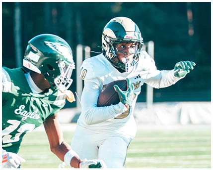 A person wearing sunglasses and a headset is speaking intensely while another person in a football helmet looks on in the background. Description generated by AI