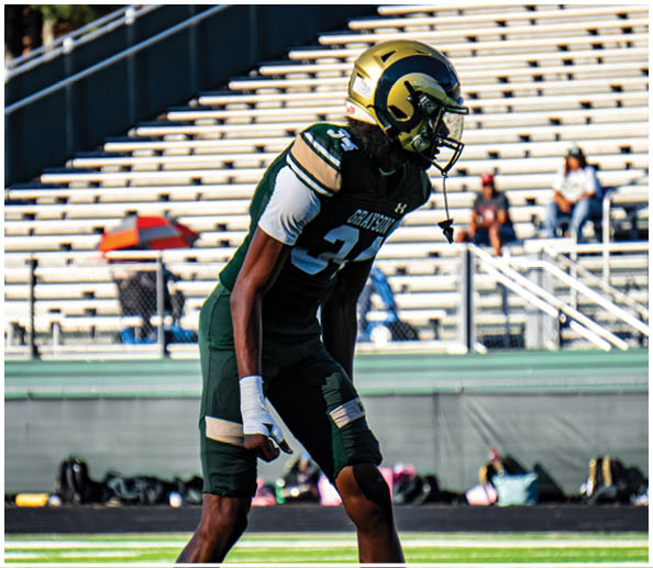 A person in a white and gold football uniform is crouched in an athletic stance on a green football field with other players and a coach in the background. Description generated by AI