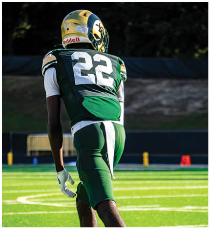 A person with long dreadlocks wearing a white football jersey with the name Grayson and the number 28 stands in front of two other people in similar football uniforms and helmets. Description generated by AI