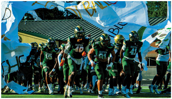 A group of football players in white uniforms and helmets run energetically through a torn banner held by cheerleaders on a field. Description generated by AI