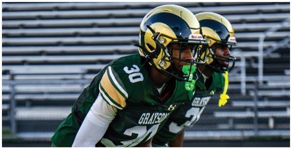 Two people wearing white sports uniforms with green and yellow stripes stand on a field with others in the background. Description generated by AI