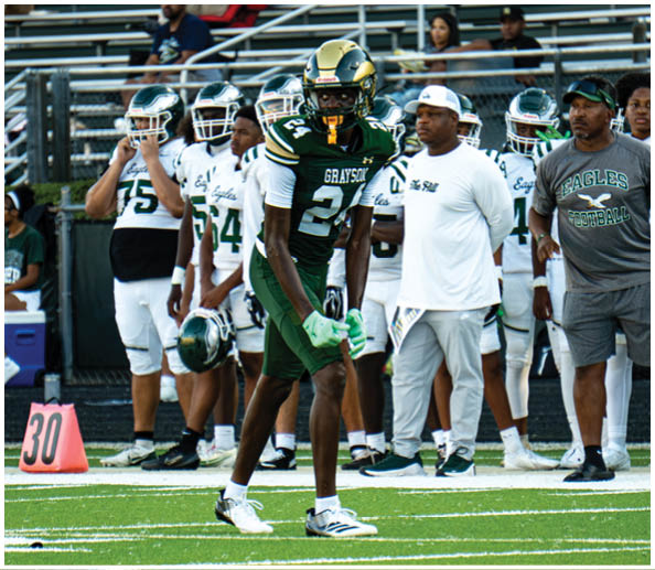 A person in a white and green football uniform with a gold helmet is on a field facing away from the camera while another person in a maroon and gold uniform stands in the background near empty bleachers. Description generated by AI
