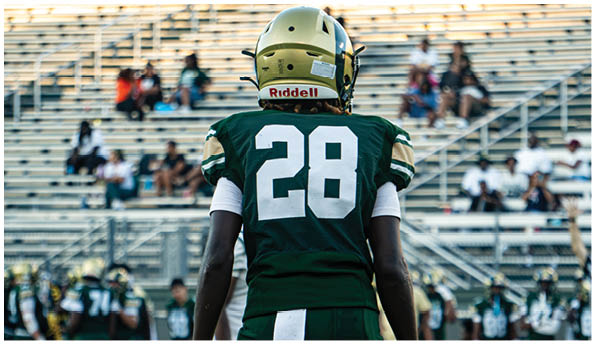 A person wearing a white football uniform and helmet is running with a football on a field while people in green cheerleading outfits stand in the background. Description generated by AI