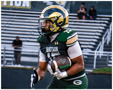 A person wearing a white football uniform with a gold and black helmet is kneeling on a green football field with arms extended forward. Description generated by AI