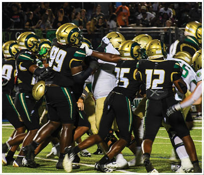 Two people in white and green football uniforms stand on a field with one clenching a fist and a referee and opposing players in red uniforms visible in the background. Description generated by AI