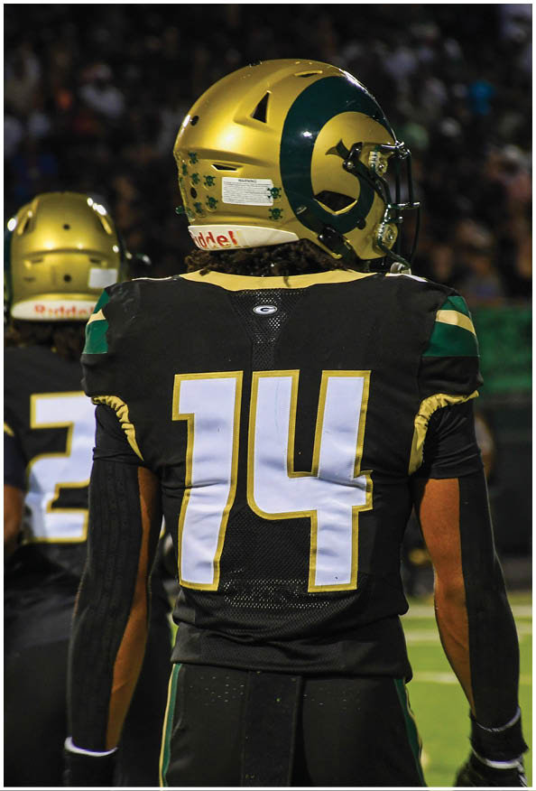 A person in a white and gold football uniform is crouched in an athletic stance on a green football field with other players and a coach in the background. Description generated by AI