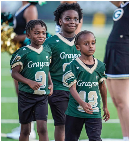 A person with long dreadlocks wearing a white football jersey with the name Grayson and the number 28 stands in front of two other people in similar football uniforms and helmets. Description generated by AI