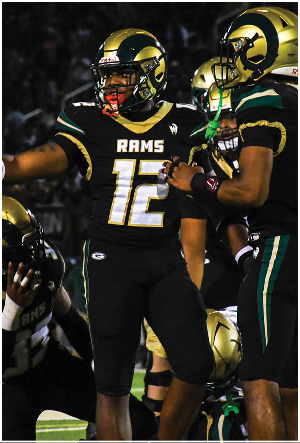A person in a white and gold football uniform is crouched in an athletic stance on a green football field with other players and a coach in the background. Description generated by AI