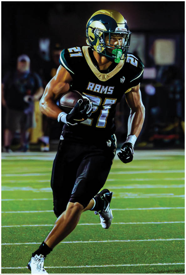 A person in a white and gold football uniform is crouched in an athletic stance on a green football field with other players and a coach in the background. Description generated by AI