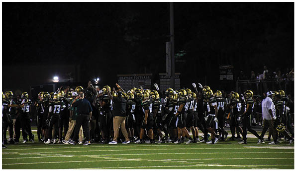 A group of football players in white uniforms and helmets run energetically through a torn banner held by cheerleaders on a field. Description generated by AI