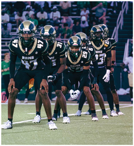 A person with long dreadlocks wearing a white football jersey with the name Grayson and the number 28 stands in front of two other people in similar football uniforms and helmets. Description generated by AI