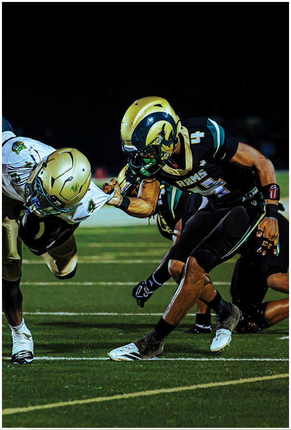A person in a white and gold football uniform is crouched in an athletic stance on a green football field with other players and a coach in the background. Description generated by AI
