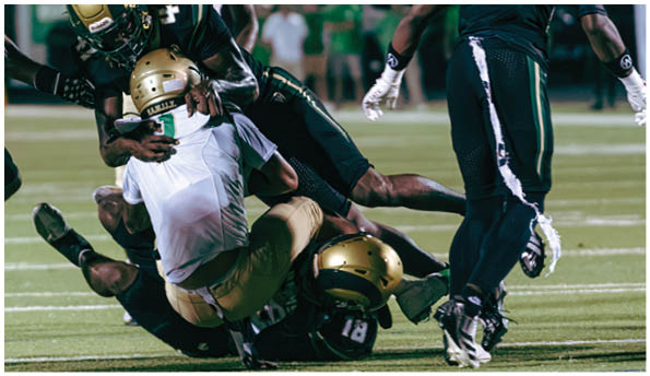 A group of football players in white uniforms and helmets run energetically through a torn banner held by cheerleaders on a field. Description generated by AI