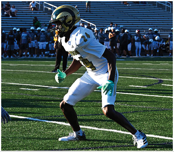 A person in a white and gold football uniform is crouched in an athletic stance on a green football field with other players and a coach in the background. Description generated by AI