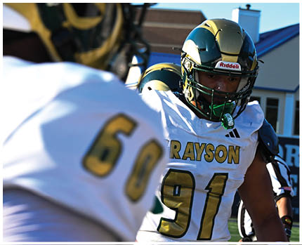 A person wearing a gold and black football helmet and a white jersey with the word “BRAVES" visible is looking to the side against a blue sky background. Description generated by AI