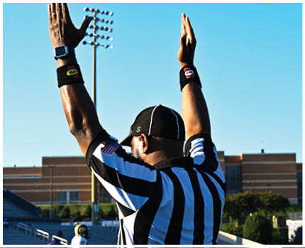 A person wearing a white and gold football uniform with the number 20 stands on a field while two other people in red and gold football uniforms are visible in the background near empty bleachers. Description generated by AI