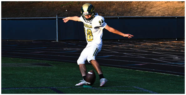 A person wearing a white sports uniform with the number 47 is standing on a field with other people in similar uniforms and spectators seated in bleachers in the background. Description generated by AI