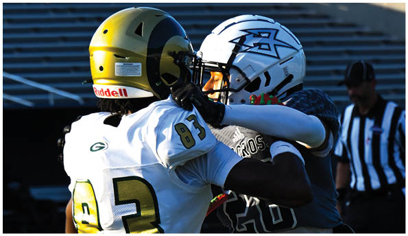 A person wearing a white football uniform and helmet is running with a football on a field while people in green cheerleading outfits stand in the background. Description generated by AI