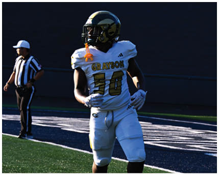 Three people wearing white and gold football uniforms and helmets are standing and interacting near a chain-link fence with trees and bleachers in the background. Description generated by AI