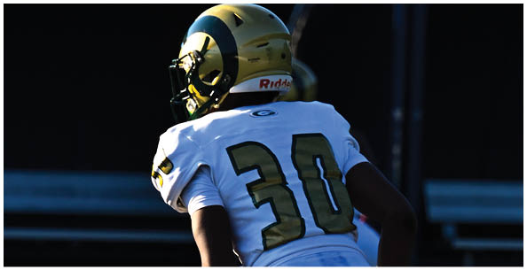 Four people wearing white football uniforms with gold helmets and large numbers on their backs are standing on a field. Description generated by AI