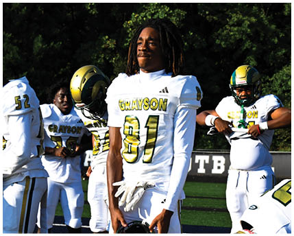 A person wearing a white football uniform with a gold and black helmet is kneeling on a green football field with arms extended forward. Description generated by AI