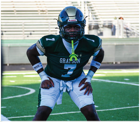 A person in a white and gold football uniform is crouched in an athletic stance on a green football field with other players and a coach in the background. Description generated by AI