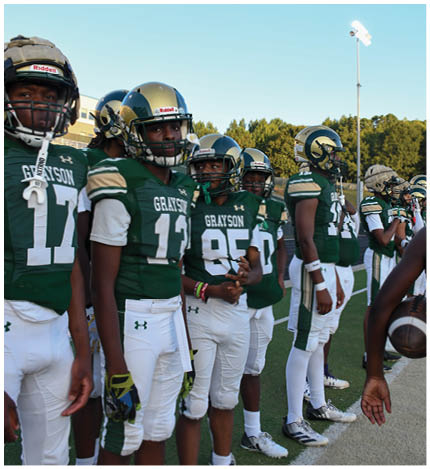 A person with long dreadlocks wearing a white football jersey with the name Grayson and the number 28 stands in front of two other people in similar football uniforms and helmets. Description generated by AI