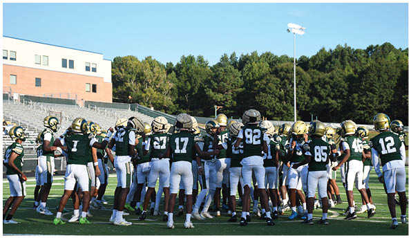 A group of football players in white uniforms and helmets run energetically through a torn banner held by cheerleaders on a field. Description generated by AI