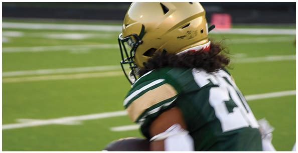 Two people wearing white sports uniforms with green and yellow stripes stand on a field with others in the background. Description generated by AI
