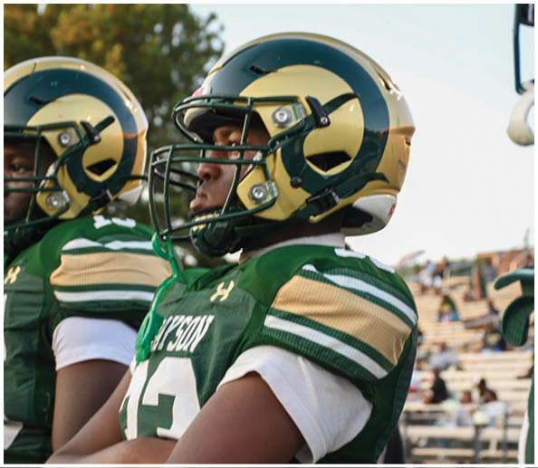 A person in a white and green football uniform with a gold helmet is on a field facing away from the camera while another person in a maroon and gold uniform stands in the background near empty bleachers. Description generated by AI